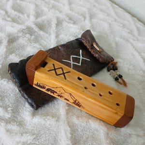 Op de afbeelding: Een houten fluit met een lichtbruine afwerking en donkerbruine accenten. De fluit heeft een gesneden berg- en berenscène, samen met zwarte geometrische symbolen. Het rust op een bruin leren zakje met witte symbolen en een kralenkwast.
