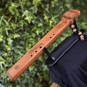 May include: A brown wooden flute featuring engraved bird and geometric symbols, resting on a black stand. The instrument has multiple finger holes, a block at the top with etched markings, and decorative wooden beads hanging from a black cord.