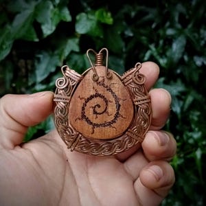Puede incluir: Un colgante con una pieza central de madera de color marrón claro en forma de lágrima con un diseño de espiral oscuro grabado. La madera está encerrada en un marco intrincado de alambre de cobre tejido con espirales decorativas.