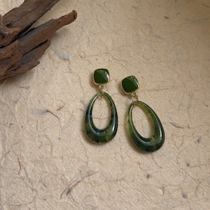May include: A pair of green drop earrings. Each earring features a square green stud connected to a gold-colored metal loop. The loop is a teardrop shape with a translucent green hue, showcasing a marbled effect. The earrings are displayed on a textured, light brown surface.