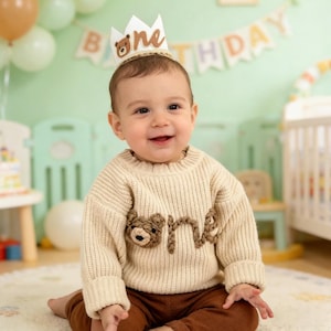 May include: A baby wearing a cream-coloured sweater with a bear design and the word "one". The baby is also wearing a white crown with a bear and the word "one". Balloons and a banner that says "Happy Birthday" are in the background.