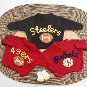 Puede incluir: Tres su&eacute;teres de beb&eacute; tejidos en negro y rojo, cada uno con dise&ntilde;os deportivos. El su&eacute;ter negro dice "Steelers" con un bal&oacute;n de f&uacute;tbol. Los su&eacute;teres rojos dicen "49ers" y "Rebels" con un bal&oacute;n de f&uacute;tbol y baloncesto.