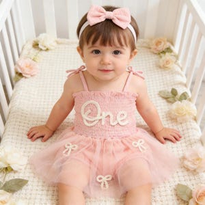 May include: A baby wearing a pink dress with the word "One" on the bodice and a matching bow headband. The dress has a tulle skirt and bow accents. The baby is sitting in a white crib.