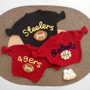 Puede incluir: Tres su&eacute;teres de punto en negro y rojo, con dise&ntilde;os deportivos. El su&eacute;ter negro tiene "Steelers" y un bal&oacute;n de f&uacute;tbol americano, mientras que los su&eacute;teres rojos muestran "49ers" con un bal&oacute;n de f&uacute;tbol y "Rebels" con una pelota de baloncesto. Tambi&eacute;n se ven peque&ntilde;os zapatos de beb&eacute; blancos y amarillos.