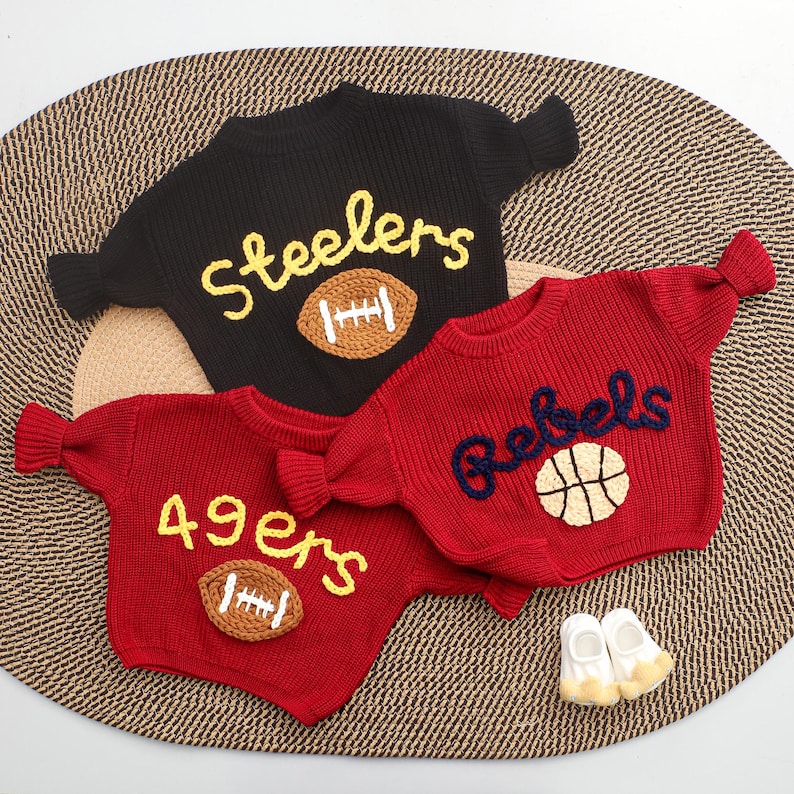Puede incluir: Tres su&eacute;teres de punto en negro y rojo, con dise&ntilde;os deportivos. El su&eacute;ter negro tiene "Steelers" y un bal&oacute;n de f&uacute;tbol americano, mientras que los su&eacute;teres rojos muestran "49ers" con un bal&oacute;n de f&uacute;tbol y "Rebels" con una pelota de baloncesto. Tambi&eacute;n se ven peque&ntilde;os zapatos de beb&eacute; blancos y amarillos.