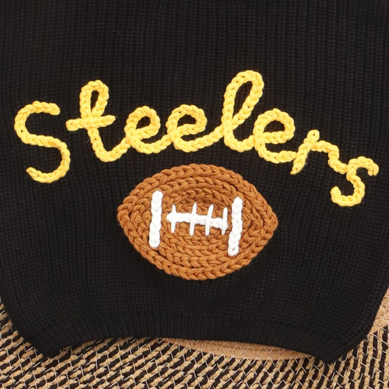 Puede incluir: Su&eacute;ter de punto negro con la palabra "Steelers" en escritura amarilla sobre un motivo de bal&oacute;n de f&uacute;tbol marr&oacute;n y blanco. El su&eacute;ter est&aacute; hecho de una tela de punto suave y texturizada.