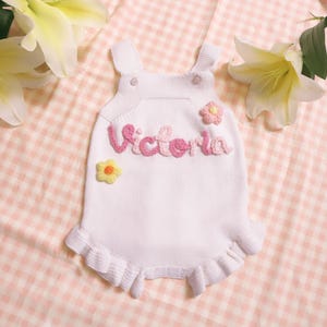 Puede incluir: Unisie de beb&eacute; de punto blanco con una flor de crochet rosa y amarilla y el nombre "Victoria" escrito en hilo rosa.