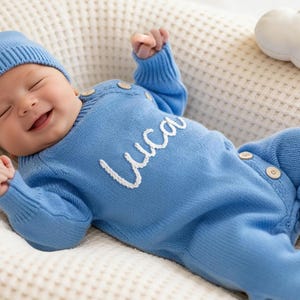 May include: A smiling baby wearing a light blue knit outfit and matching hat with a pom-pom. The outfit has the name "Luca" embroidered in white. The baby is lying on a cream-colored surface.