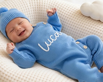 Puede incluir: Un bebé sonriente con un conjunto de punto azul claro y un gorro a juego con pompón. El nombre "Luca" está bordado en blanco en el conjunto. El bebé está acostado sobre una superficie color crema.