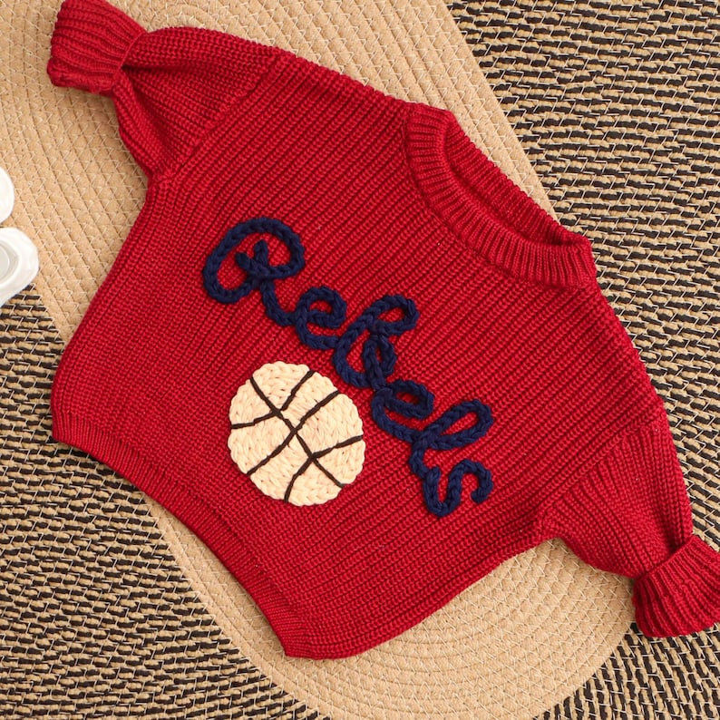 Puede incluir: Un su&eacute;ter rojo tejido con la palabra "Rebels" en escritura azul marino y un gr&aacute;fico de baloncesto. El su&eacute;ter tiene cuello redondo y mangas largas con pu&ntilde;os acanalados. El su&eacute;ter est&aacute; hecho de un tejido grueso y texturizado.