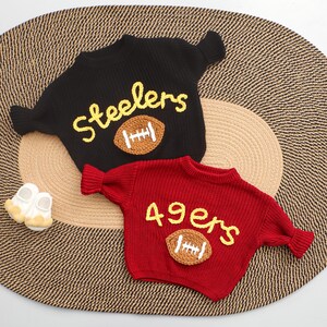 Puede incluir: Dos su&eacute;teres de punto, uno negro y otro rojo, con dise&ntilde;os de f&uacute;tbol americano. El su&eacute;ter negro tiene "Steelers" en letras amarillas y una aplicaci&oacute;n de bal&oacute;n de f&uacute;tbol marr&oacute;n. El su&eacute;ter rojo presenta "49ers".