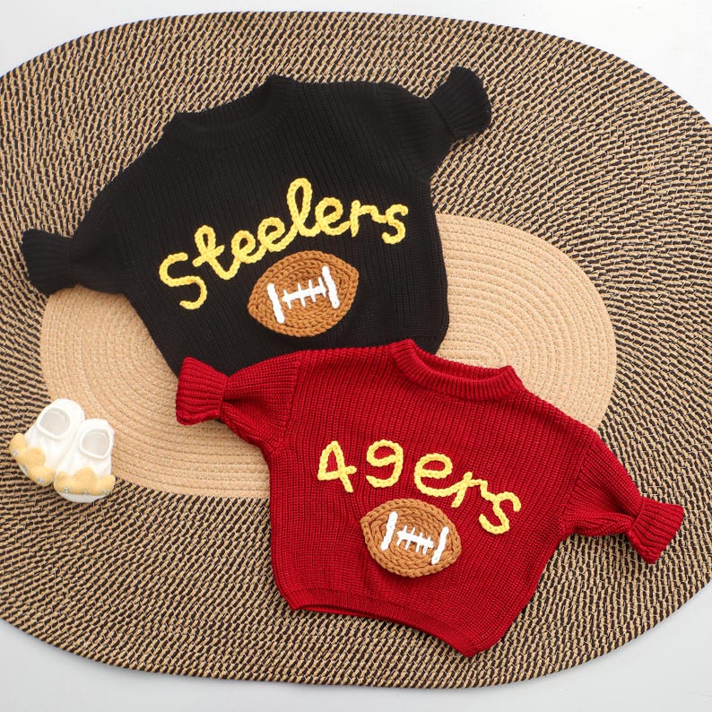 Puede incluir: Dos su&eacute;teres de punto, uno negro y otro rojo, con dise&ntilde;os de f&uacute;tbol americano. El su&eacute;ter negro tiene "Steelers" en letras amarillas y una aplicaci&oacute;n de bal&oacute;n de f&uacute;tbol marr&oacute;n. El su&eacute;ter rojo presenta "49ers".