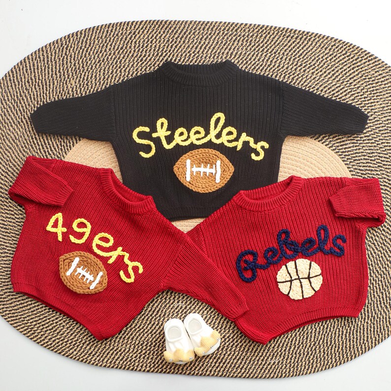 Puede incluir: Tres su&eacute;teres de beb&eacute; tejidos en negro y rojo, cada uno con dise&ntilde;os deportivos. El su&eacute;ter negro dice "Steelers" con un bal&oacute;n de f&uacute;tbol. Los su&eacute;teres rojos dicen "49ers" y "Rebels" con un bal&oacute;n de f&uacute;tbol y baloncesto.