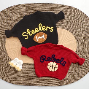 Puede incluir: Dos su&eacute;teres de punto, uno negro con "Steelers" y un bal&oacute;n de f&uacute;tbol americano, el otro rojo con "Rebels" y una pelota de baloncesto. Peque&ntilde;os zapatos de beb&eacute; blancos y amarillos est&aacute;n junto a los su&eacute;teres. Los su&eacute;teres est&aacute;n sobre una estera tejida.