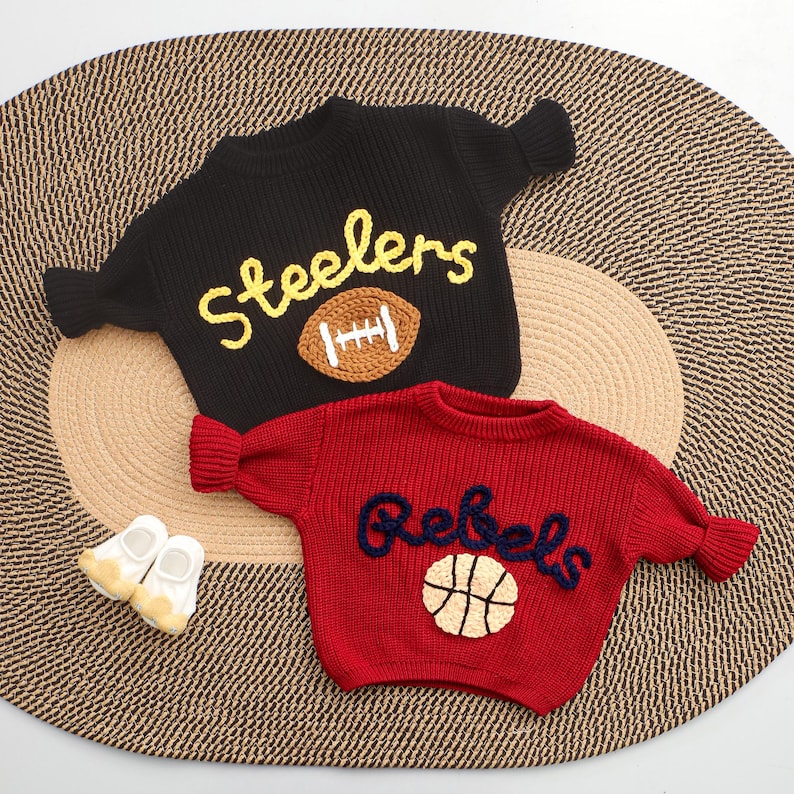 Puede incluir: Dos su&eacute;teres de punto, uno negro con "Steelers" y un bal&oacute;n de f&uacute;tbol americano, el otro rojo con "Rebels" y una pelota de baloncesto. Peque&ntilde;os zapatos de beb&eacute; blancos y amarillos est&aacute;n junto a los su&eacute;teres. Los su&eacute;teres est&aacute;n sobre una estera tejida.