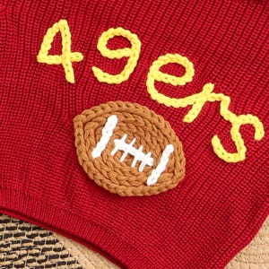 Puede incluir: Tejido de punto rojo con texto bordado amarillo "49ers" y una aplicaci&oacute;n de f&uacute;tbol marr&oacute;n y blanco. El tejido tiene un aspecto texturizado.