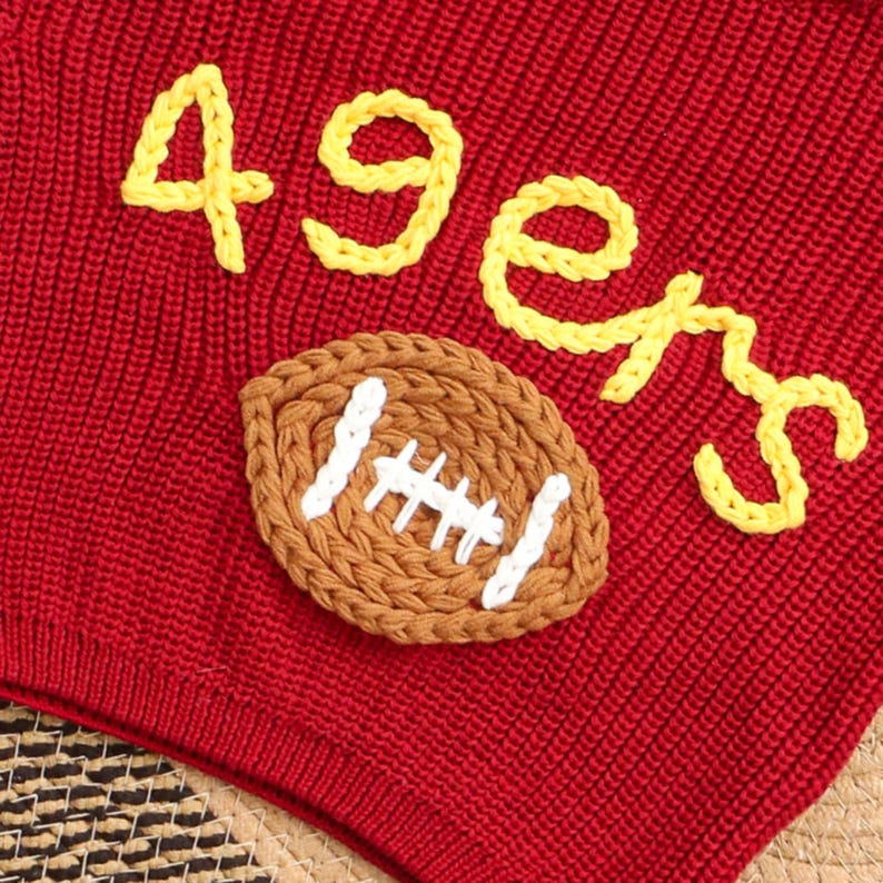 Puede incluir: Tejido de punto rojo con texto bordado amarillo "49ers" y una aplicaci&oacute;n de f&uacute;tbol marr&oacute;n y blanco. El tejido tiene un aspecto texturizado.
