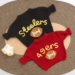 Puede incluir: Dos su&eacute;teres de beb&eacute; tejidos, uno negro con "Steelers" en amarillo y un bal&oacute;n de f&uacute;tbol, el otro rojo con "49ers" y un bal&oacute;n de f&uacute;tbol. Los su&eacute;teres est&aacute;n sobre una estera tejida, con un par de zapatos blancos.