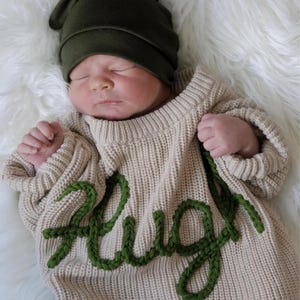 May include: A newborn baby wearing a beige knit sweater with the word "Hug" in green yarn. The baby is also wearing a dark green beanie. The baby is lying on a white, fluffy surface.