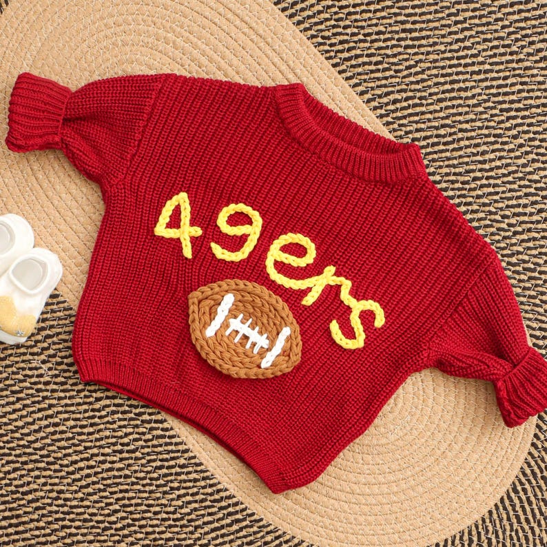 Puede incluir: Un su&eacute;ter rojo tejido con la palabra "49ers" en letras amarillas y una aplicaci&oacute;n de f&uacute;tbol americano marr&oacute;n y blanco. El su&eacute;ter tiene mangas largas y cuello redondo. El su&eacute;ter se muestra sobre una estera tejida.