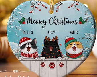 May include: A heart-shaped Christmas ornament with a winter scene featuring three cats wearing Santa hats and scarves. The ornament says "Meowy Christmas" and has the names Bella, Lucy, and Milo.