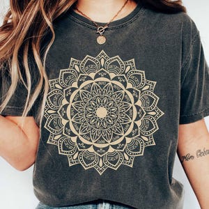 May include: A dark gray t-shirt with a large, intricate gold mandala design. The mandala features a repeating pattern of geometric shapes and the word "RESEST" written around the outer edge.
