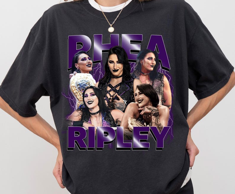 Puede incluir: Camiseta negra con un gr&aacute;fico morado de Rhea Ripley y el texto "RHEA RIPLEY". El dise&ntilde;o incluye im&aacute;genes de Rhea Ripley con un efecto de rayo p&uacute;rpura. La camiseta tiene cuello redondo y mangas cortas.