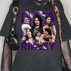 Puede incluir: Camiseta gris oscuro con un gr&aacute;fico de Rhea Ripley y el texto "Rhea Ripley" en morado. El gr&aacute;fico incluye m&uacute;ltiples im&aacute;genes de Rhea Ripley con un dise&ntilde;o de rayo morado.