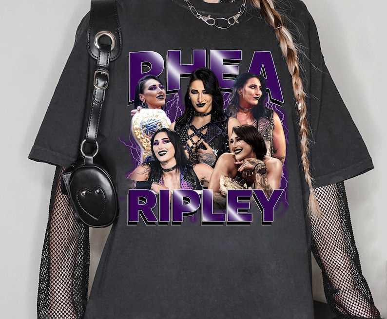 Puede incluir: Camiseta gris oscuro con un gr&aacute;fico de Rhea Ripley y el texto "Rhea Ripley" en morado. El gr&aacute;fico incluye m&uacute;ltiples im&aacute;genes de Rhea Ripley con un dise&ntilde;o de rayo morado.