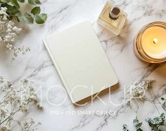 May include: A white blank notebook on a marble surface with a lit candle, a bottle of perfume, and white flowers.