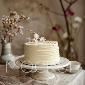 Könnte beinhalten: Ein weißer Kuchen mit weißem Frosting und zarten rosa und weißen Blüten darauf. Der Kuchen steht auf einem weißen Kuchenständer und einer weißen Tischdecke.