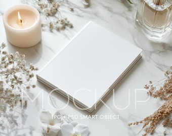 May include: A white blank book mockup on a marble surface with a lit candle, dried flowers, and a clear glass bottle of perfume.
