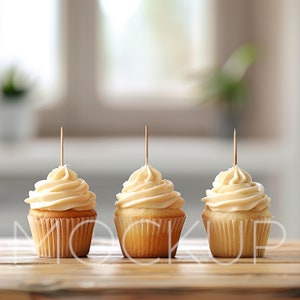 Könnte beinhalten: Drei Vanille-Cupcakes mit weißem Frosting und einem Zahnstocher in jedem. Die Cupcakes stehen auf einem Holztisch.