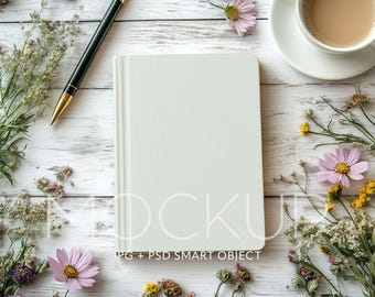 May include: A white blank notebook with a black pen on a white wooden surface with pink and yellow flowers. The text "Mockup" is printed on the notebook. The text "JPG + PSD SMART OBJECT" is printed below the notebook.