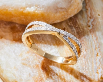 May include: A gold ring with a crisscross design, encrusted with small, sparkling diamonds. The ring is set against a neutral, textured background. The band is marked with "14K".