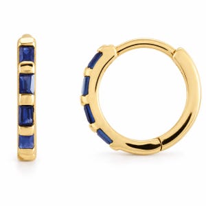 May include: A pair of gold hoop earrings featuring a row of four rectangular, deep blue sapphire gemstones set along the front curve of each hoop. The earrings have a polished gold finish and a hinged closure mechanism for secure wear.
