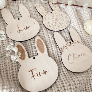 Könnte beinhalten: Osterhasen-förmige Holzornamente mit eingravierten Namen. Die hellbraunen Dekorationen weisen florale Muster und Namen wie "Lio" und "Finn" auf. Jedes Ornament hat ein kleines Loch zum Aufhängen.