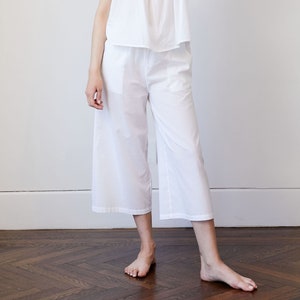 May include: White cotton pajama set. The set includes a camisole top with tie straps and wide-leg cropped pants. The model is wearing the set in a well-lit room with a white wall and a wooden floor.