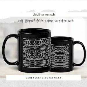 Könnte beinhalten: Zwei schwarze Keramikbecher mit weißen geometrischen Mustern. Die Becher tragen die weißen Aufschriften "Lieblingsmensch", "... weil Superheld:in schon vergeben war." und "VERSTECKTE BOTSCHAFT".