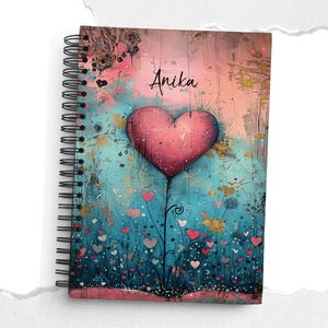 Könnte beinhalten: Spiralgebundenes Notizbuch mit einem farbenfrohen, künstlerischen Einband. Der Einband zeigt ein großes rosa Herz, kleinere Herzen und florale Elemente vor einem türkis-rosa Hintergrund. Der Name "Anika" ist auf dem Einband geschrieben.