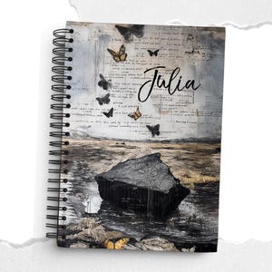 Puede incluir: Cuaderno de espiral con una cubierta de arte mixto. La cubierta presenta un paisaje con una gran formación rocosa negra, mariposas y texto manuscrito. El nombre "Julia" está escrito en cursiva. El cuaderno tiene una encuadernación en espiral negra.
