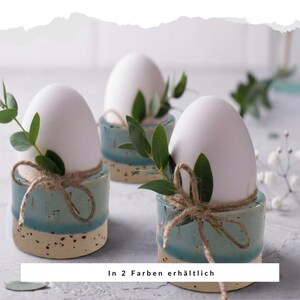 Puede incluir: Tres hueveras de cerámica en un diseño moteado azul claro y beige, cada una con un huevo blanco y ramitas verdes. Las hueveras están atadas con hilo. El texto en la parte inferior dice "In 2 Farben erhältlich".