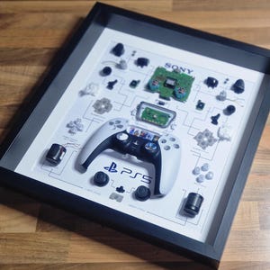 May include: A white PlayStation 5 controller disassembled and displayed in a black frame. The controller is surrounded by various small parts, including buttons, triggers, and a circuit board. The frame is on a wooden surface.