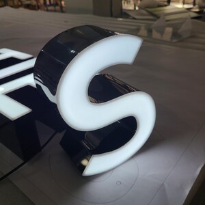 以下が含まれることがあります： 光る文字「S」のクローズアップ。 立体的な文字は、光沢のある黒い外観と明るい白い光る内部を持っています。 画像は、デザインと色のコントラストを強調しています。
