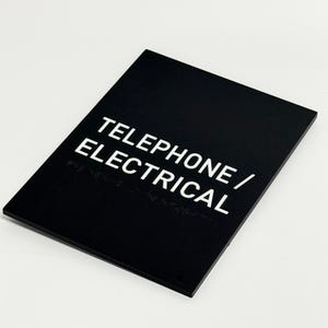 May include: A black rectangular sign with the words "TELEPHONE / ELECTRICAL" in white, sans-serif font. The sign also features Braille text. The sign is angled slightly, with a white background.
