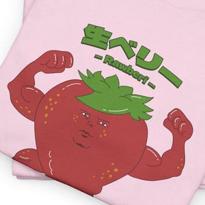 Puede incluir: Camiseta rosa con una ilustración de dibujos animados de una fresa roja con una corona de hojas verdes y un cuerpo musculoso. La fresa tiene una cara humana y está flexionando sus bíceps. El texto "Strawberry - Rawber! -" está escrito en japonés e inglés en la camiseta.