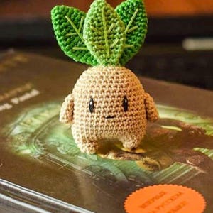 以下が含まれることがあります： タン色の体、小さな腕、緑の葉を持つ、マンドレイク植物に似たかぎ針編みの人形。人形は笑顔で、本の上に置かれています。「PDF PATTERN」の文字が見えます。