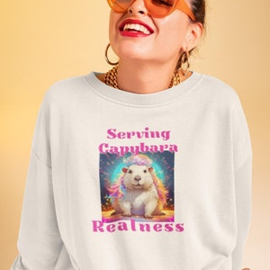 Op de afbeelding: Witte sweatshirt met een afbeelding van een capibara met kleurrijk haar en de tekst "Serving Capybara Realness". De persoon draagt een oranje zonnebril en gouden sieraden. De achtergrond is een gele tot oranje gradiënt.