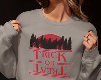 May include: Gray sweatshirt with a red graphic featuring a forest silhouette, a red sun, and the words "TRICK OR TREAT" in red. The design includes silhouettes of people riding bicycles. A casual, festive garment.