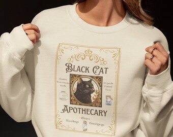 May include: A cream-coloured sweatshirt with a vintage-style graphic. The graphic reads "Black Cat Apothecary" with a black cat illustration. The design includes decorative borders and apothecary-style elements, creating a whimsical, gothic aesthetic.